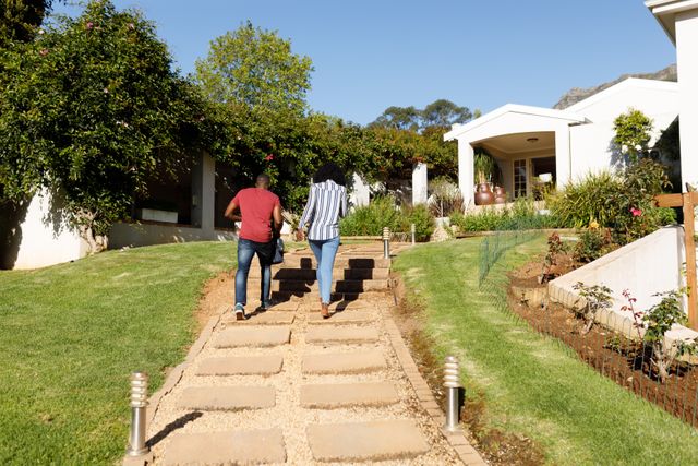 This image captures an African-American couple walking up a path towards their home, surrounded by a well-maintained lawn and various plants. Ideal for use in real estate, home improvement, gardening, and lifestyle content to depict family life, homeownership, and outdoor living.