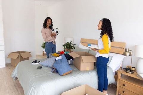 Mother and teenage daughter unpacking boxes in sunlit bedroom holding soccer ball