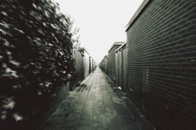 Narrow urban alleyway between buildings in black and white, creating a dramatic and intense feel. Appears as a vanishing point perspective with a close-up view of brick and metal structures. Suitable for editorial use illustrating urban environments, documentaries, and industrial architectural subjects. Perfect for backgrounds in graphic design projects.