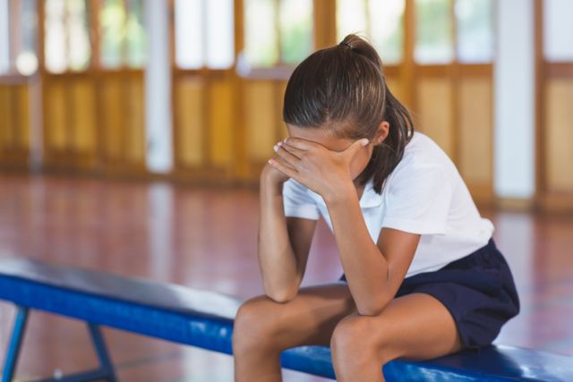 Young girl sitting alone on a bench in a school gym, covering her face with her hands. The image conveys emotions of sadness, stress, and solitude. Useful for illustrating concepts related to mental health, childhood emotions, school life, and the importance of emotional well-being in educational settings.