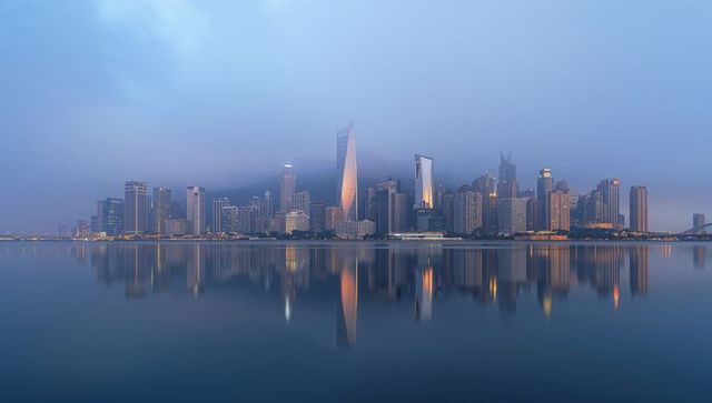 Mist-cloaked skyline reflecting in glassy bay at dusk with illuminated landmark towers