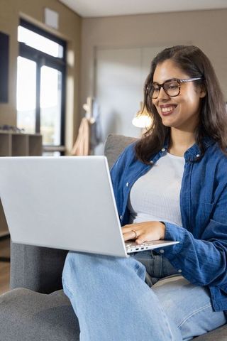 Young woman working from home on laptop, smiling, casual denim, glasses, cozy modern living room