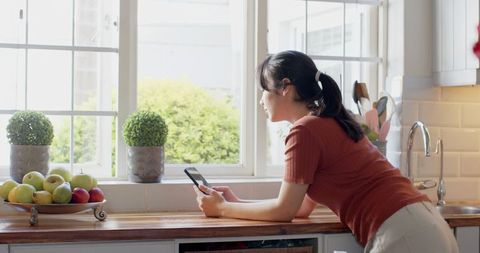 Asian Woman Relaxing in Modern Kitchen with Smartphone and Fruit Tray