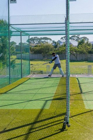 Cricket batsman practicing in net cage driving shot on turf during sunny suburban training