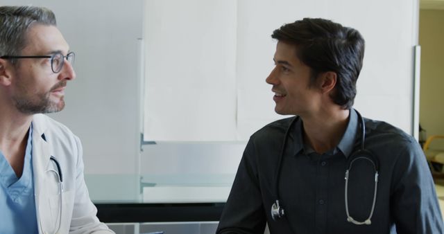 Two male doctors are sitting and discussing a medical case in a hospital setting. Both appear engaged and focused, wearing stethoscopes. Suitable for themes related to healthcare, medical teamwork, professional collaboration, and patient care.