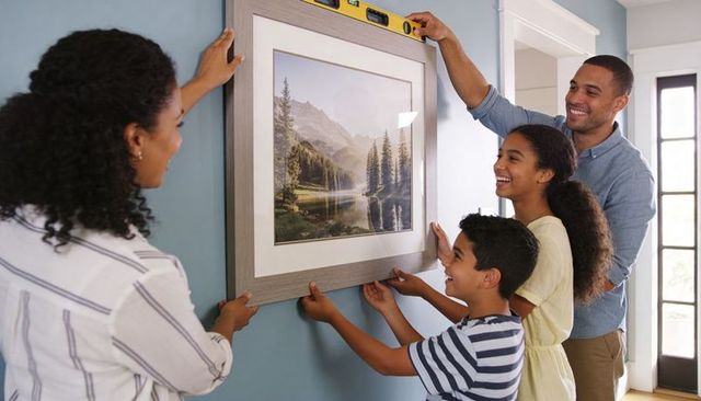 Family hanging framed mountain landscape on blue wall using spirit level, smiling together in entryw
