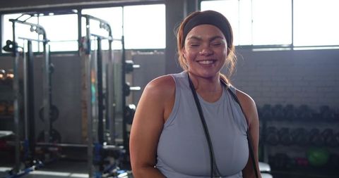 Smiling woman wearing headband and holding jump rope after workout in bright gym interior