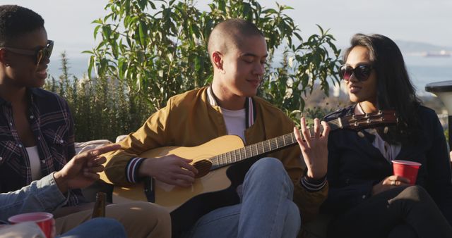 Group of friends socializing outdoors with one playing guitar and others enjoying. Used in contexts illustrating friendship, social gatherings, music, leisure activities, and youthful lifestyles. Ideal for promoting social events, outdoor activities, or music-related products and services.