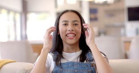 Cheerful Teenage Girl Sitting on Couch at Home in Denim Overalls