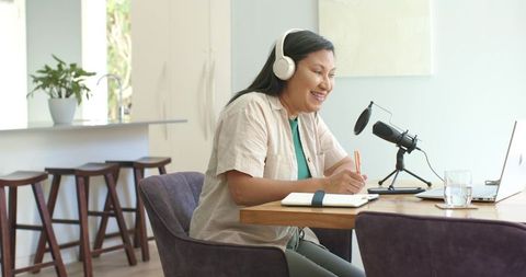 Middle-aged woman recording podcast in sunlit modern kitchen workspace with laptop