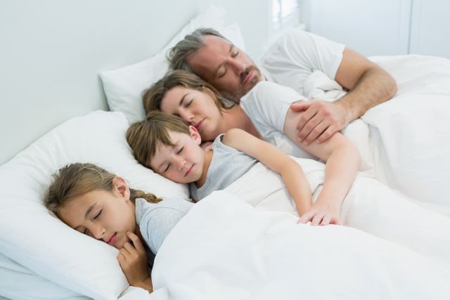 Family of four sleeping together in bed, showing a peaceful and loving moment. Ideal for use in advertisements for bedding, family-oriented products, or articles about family bonding and sleep health.