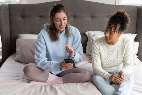 Two women sharing smartphone while lounging on bed, cozy loungewear and conversation
