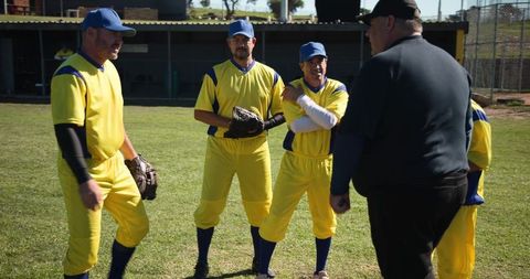 Umpire briefing yellow-uniform baseball players listening on sunlit grass field before game