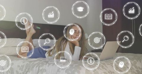 Young woman lying on bed watching laptop with floating education and tech icons