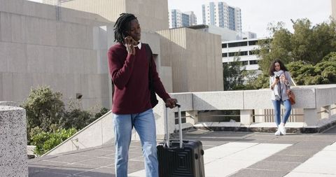 African American man talking on phone pulling suitcase across urban plaza