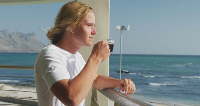 Young blond man standing on oceanfront balcony, enjoying coffee on a sunny day. Ocean and mountains visible in the background. Suitable for topics related to relaxation, vacation, leisure activities, morning routines, and summertime enjoyment.