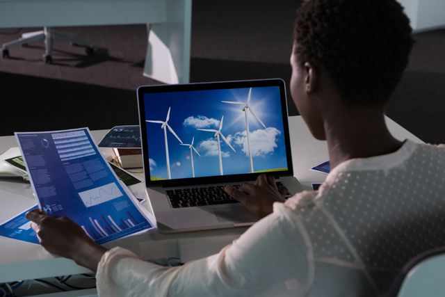 Female executive analyzing renewable energy data on laptop in modern office. She is holding charts and graphs, indicating a focus on sustainability and eco-friendly business practices. Ideal for use in articles or presentations about renewable energy, business innovation, and professional work environments.