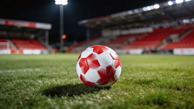 Resting red-and-white soccer ball on stadium pitch under floodlights at night with empty red stands