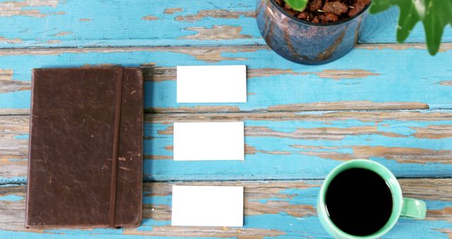 A vintage brown notebook lies next to blank business cards and a green cup of coffee on a rustic blue wooden table, with copy space. The arrangement suggests a creative brainstorming session or a casual business meeting setup.