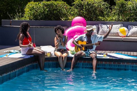 African american friends relaxing at backyard poolside, dipping feet, playing guitar