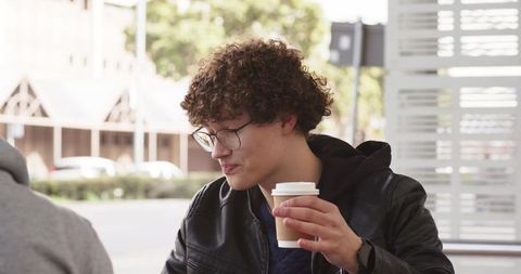 Curly-haired man wearing glasses and leather jacket holding coffee cup on urban cafe patio