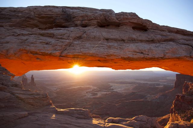 This image captures a stunning sunrise through Mesa Arch in Canyonlands National Park, Utah. The glowing sun illuminates the natural rock arch and casts a warm light across the surrounding desert landscape. Perfect for travel brochures, adventure blogs, nature documentaries, and outdoor photography collections highlighting national parks and scenic formations.