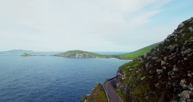 Beautiful coastal road winding along rocky cliffs with a stunning view of the blue sea and green hills. Ideal for use in travel blogs, tourism promotions, landscape photography displays, and outdoor adventure advertisements.