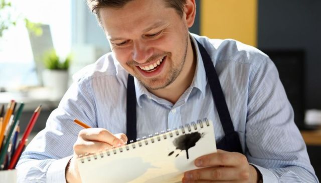 Smiling male illustrator sketching spiral sketchbook, wearing apron at bright studio desk