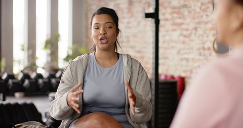 Two women discussing fitness goals while coaching in sunlit gym with dumbbells and plants