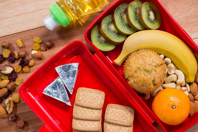 This image shows a healthy lunchbox filled with a variety of nutritious foods including kiwi slices, a banana, an orange, a muffin, biscuits, cheese, and a mix of nuts and dried fruits. A bottle of oil is also present. Ideal for illustrating balanced diet concepts, school lunch ideas, meal prep, and healthy eating habits.