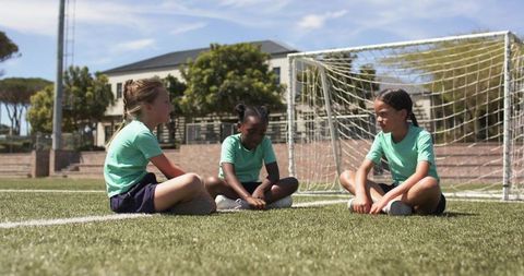 Three african american teen girls sitting on soccer field turf by goalpost in uniforms