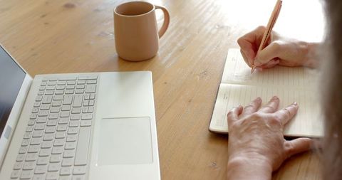 Mature Woman Writing in Notebook at Wooden Desk with Laptop and Coffee