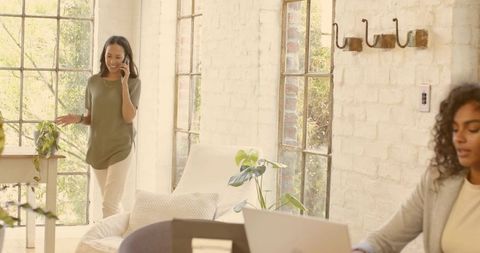 Woman talking on smartphone walking through sunlit loft office with plants and coworking vibe
