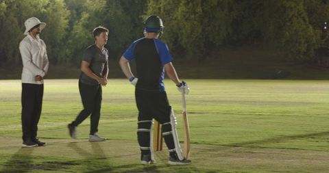 Night cricket match batsman standing at crease under floodlights with bowler and umpire