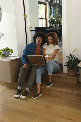 Couple sitting on steps unpacking boxes holding framed artwork and coffee in cozy entryway