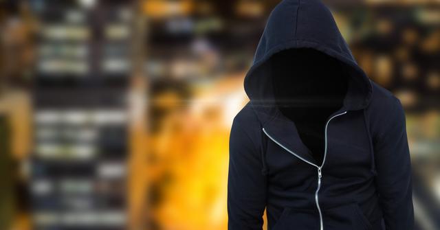 Hooded figure silhouetted with no visible face stands in front of city night lights. Suitable for illustrating themes of anonymity, cybercrime, mystery, urban life, and security. Great usage in crime documentaries, security software visuals, or fiction covers.