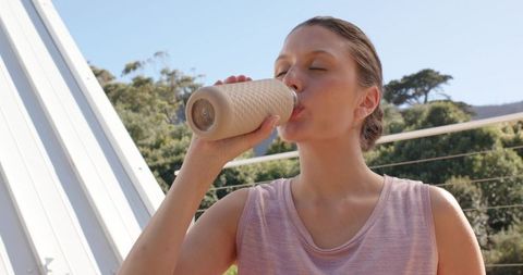 African american woman drinking from insulated bottle on sunlit balcony, summer wellness
