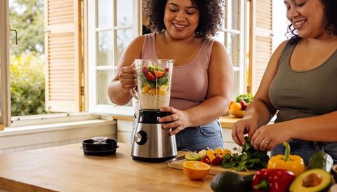 Smiling women blending green smoothie while chopping fresh fruit and leafy greens in sunny kitchen