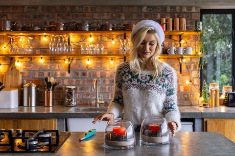 Woman in santa hat placing glass candle centerpieces on rustic kitchen countertop with string lights
