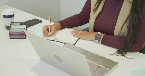 Mid adult woman writing notes at laptop in modern office workspace with smartwatch