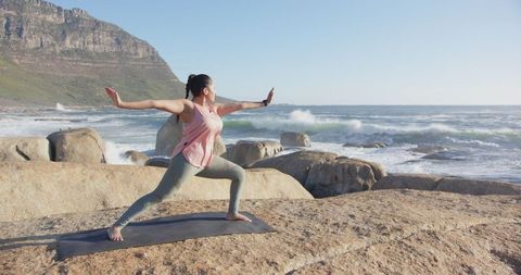 Woman practicing yoga warrior pose by ocean waves at rocky shore