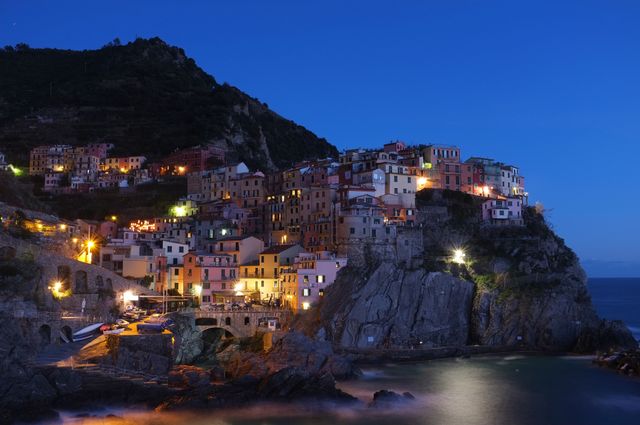 Manarola village illuminated at dusk on cliffside by Mediterranean Sea. Colorful houses light up beautifully against the twilight sky and rugged terrain. Perfect for content related to travel, Italian destinations, Mediterranean landscapes, scenic views, and architectural beauty. Useful in promotional materials for tours, vacation packages, or cultural articles.