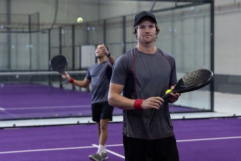 Padel players practicing on purple indoor court holding black paddle and yellow ball