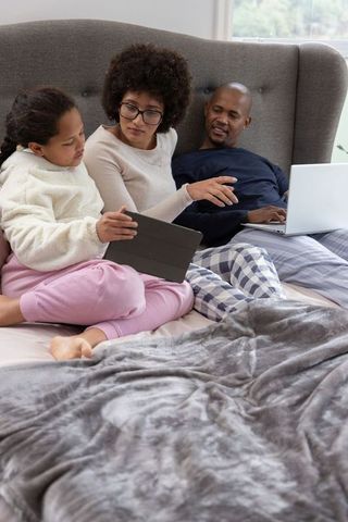 Diverse family relaxing in bed, mother pointing at tablet while father using laptop