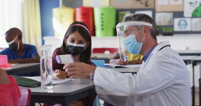 Diverse male teacher showing schoolgirl how to measure temperature, all wearing face masks. children in primary school during coronavirus covid 19 pandemic.