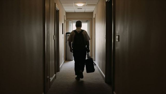 Backlit traveler walking down dim hotel corridor carrying backpack and duffel bag