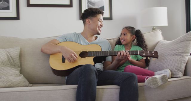 Father and daughter sit on a cozy sofa in living room, father playing guitar while smiling at daughter. Ideal for content on family bonding, music education, parenting tips, and home activities. Suitable for websites, blogs, advertisements, and social media posts focusing on family values and happy home environments.