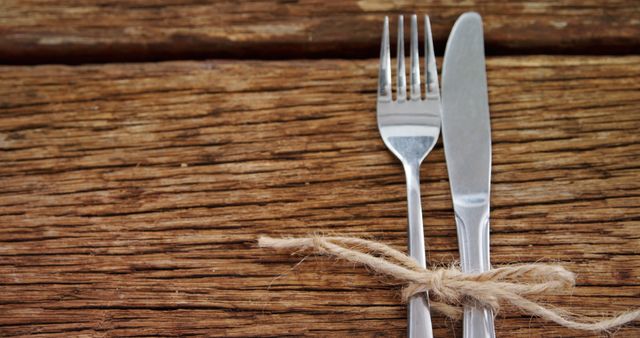 A fork and knife are tied together with a piece of twine on a rustic wooden table, with copy space. This setup suggests a cozy, homemade meal or a country-style dining experience.