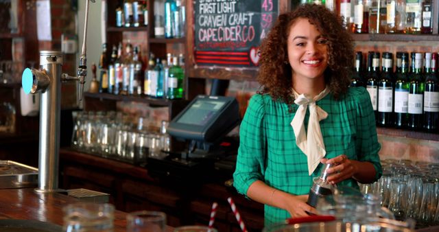 Female bartender with curly hair smiling while preparing a drink in a craft beer pub. Ideal for use in articles about hospitality industry, customer service, or promoting pubs and bars. Can be used for content related to bartending tips, drink recipes, or highlighting diverse work environments.