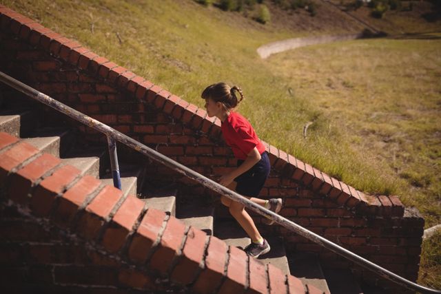 Young girl running up brick stairs during an outdoor obstacle course in a boot camp. She is wearing a red shirt and black shorts, showing determination and focus. This image can be used for promoting youth fitness programs, sports training, health and wellness campaigns, or motivational content.
